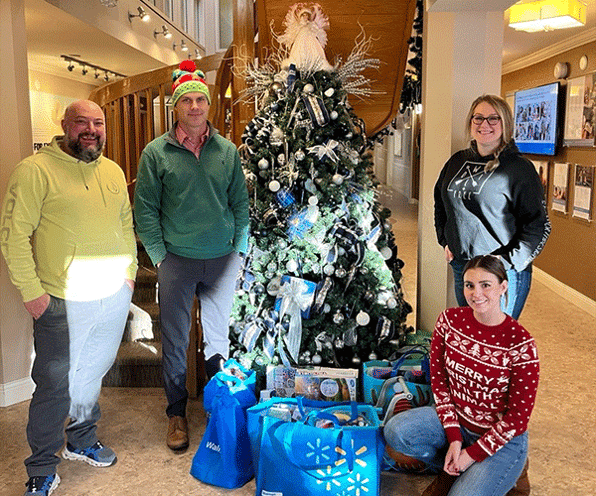 4 smiling Allnorth team members - 3 standing 1 kneeling - around a Christmas tree with blue bags under the tree.